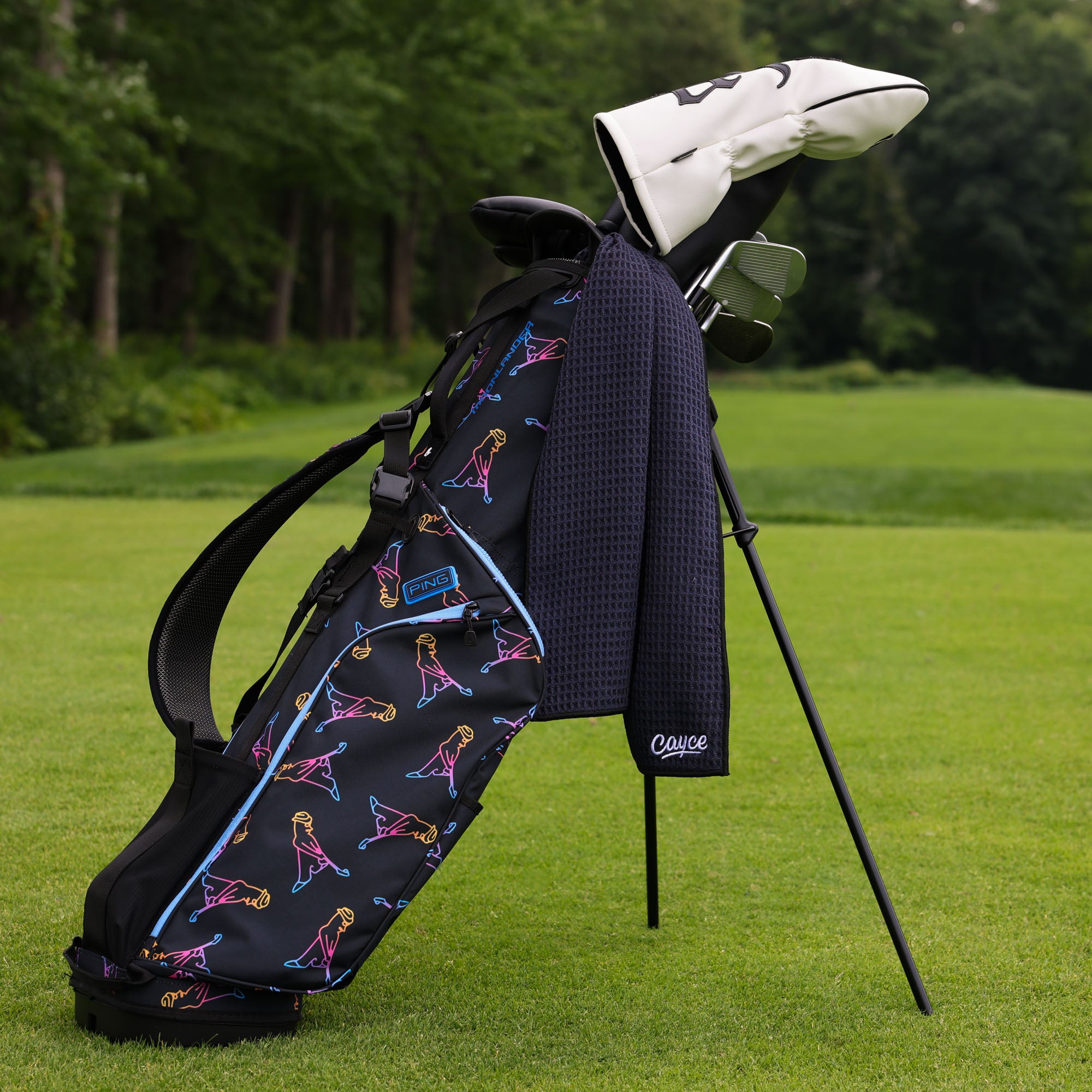A golf bag with a black base and colorful pattern print, standing on a golf course with trees in the background. A white headcover, club, and a black waffle towel are resting on the bag.