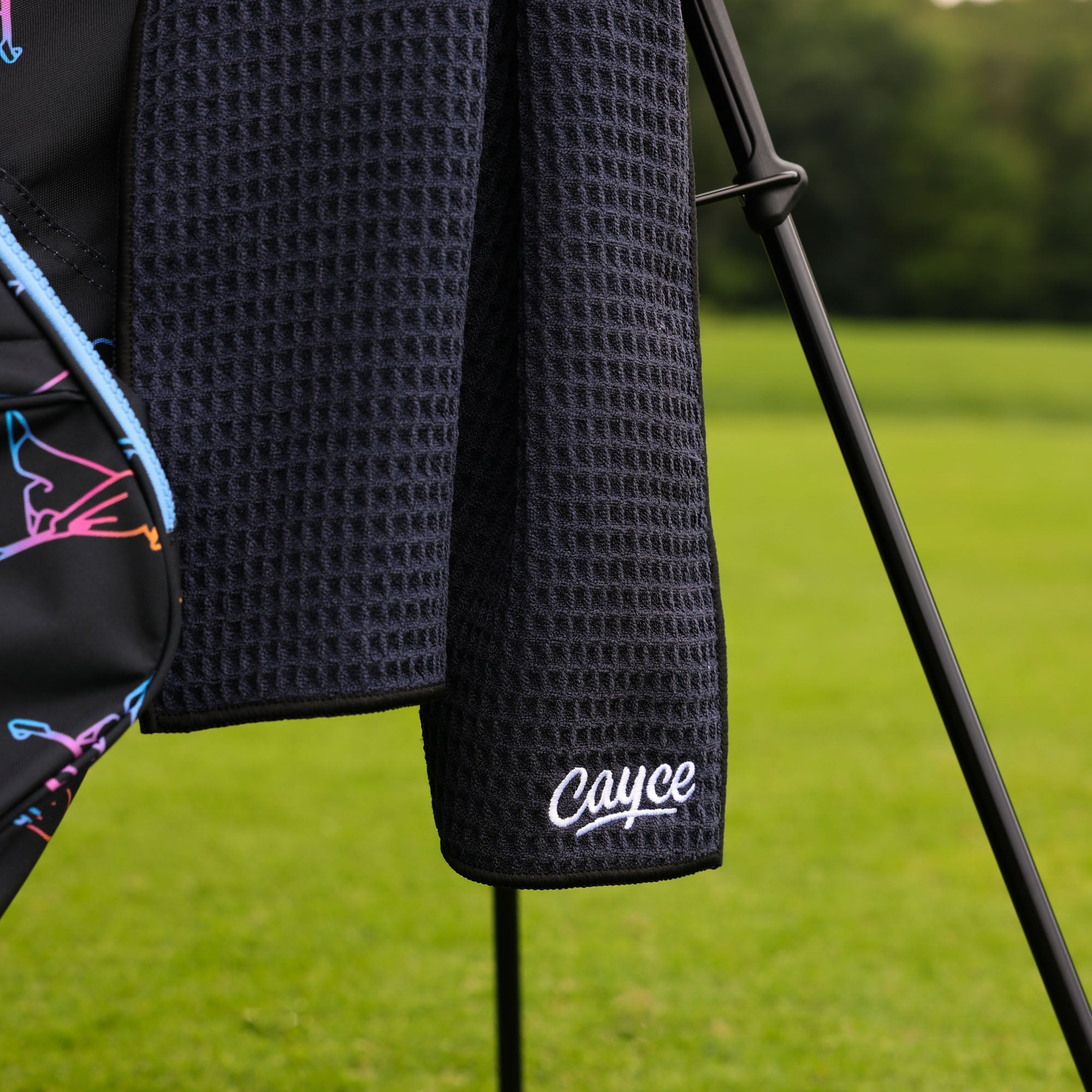 A close-up of a black waffle-weave golf towel hanging from a golf bag on a golf course, with the name 'Cayce' embroidered in white script near the bottom.