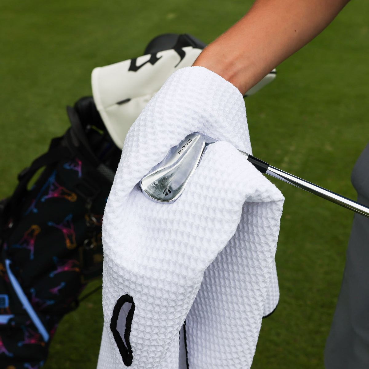 A close-up of a white waffle-textured golf towel being held by a person on a golf course, cleaning a golf club and a patterned golf bag in the background.