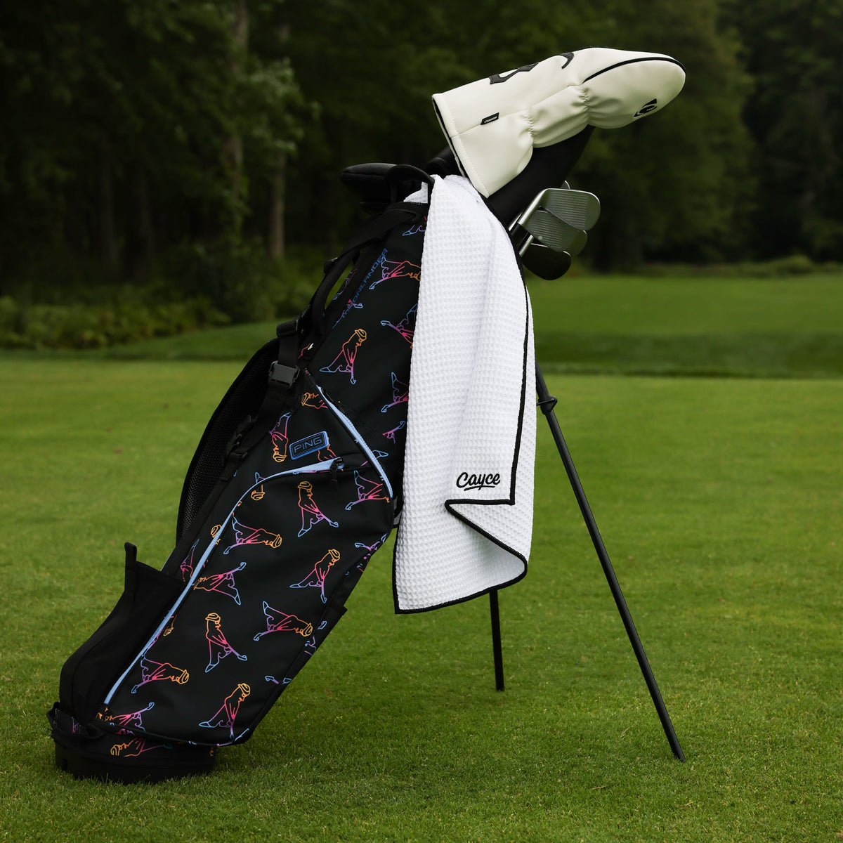 A black golf bag with a colorful pattern and a white waffle towel draped over the side, standing on a golf stand on a green grass tee box with trees in the background. The towel has the brand name 'Cayce' embroidered on it.