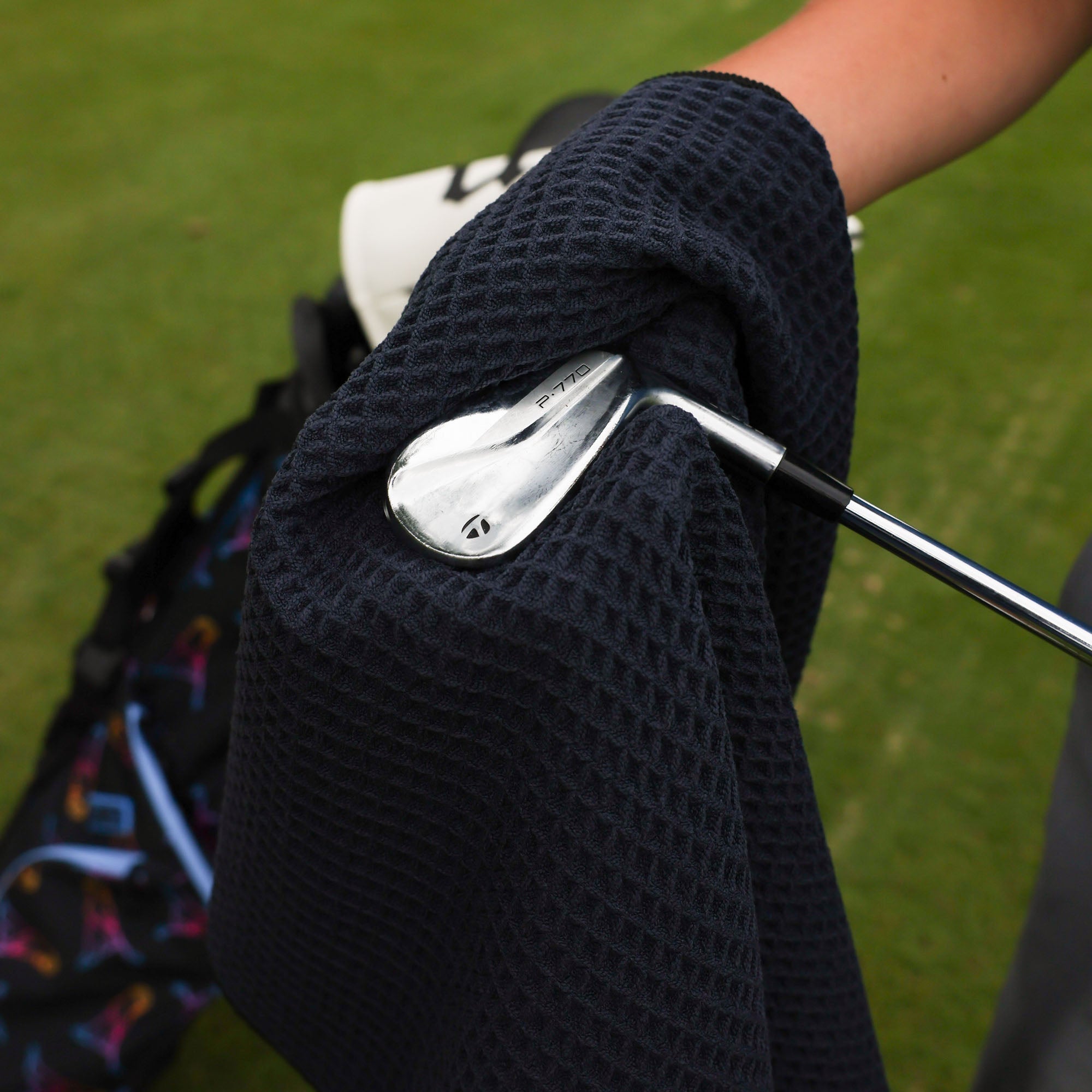 A close-up of a black waffle-weave golf towel being held by a person cleaning their club on the golf course.