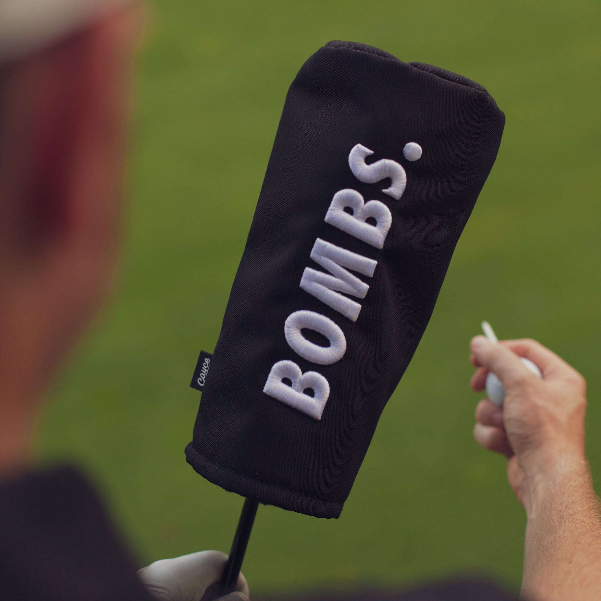 over the shoulder picture of a golfer holding a black driver head cover with 3D puff embroidered text "BOMBS." in white