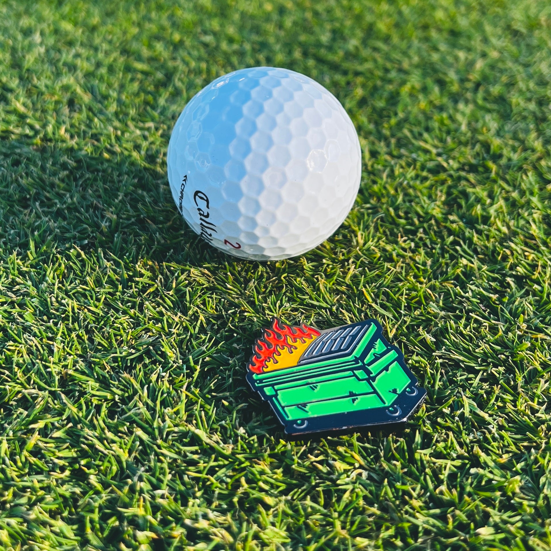 a dumpster fire golf ball marker sitting on the ground behind a golf ball on the green