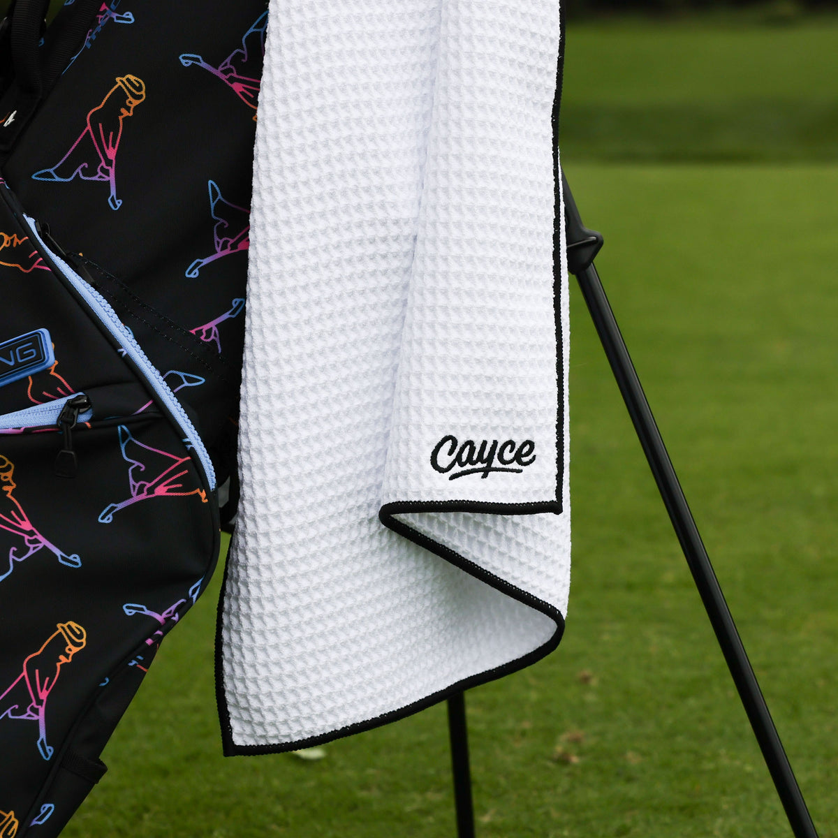 A white waffle-weave golf towel with black trim and the name 'Cayce' embroidered in black script, draped over a black golf bag with neon pattern set against a blurred green grass background.