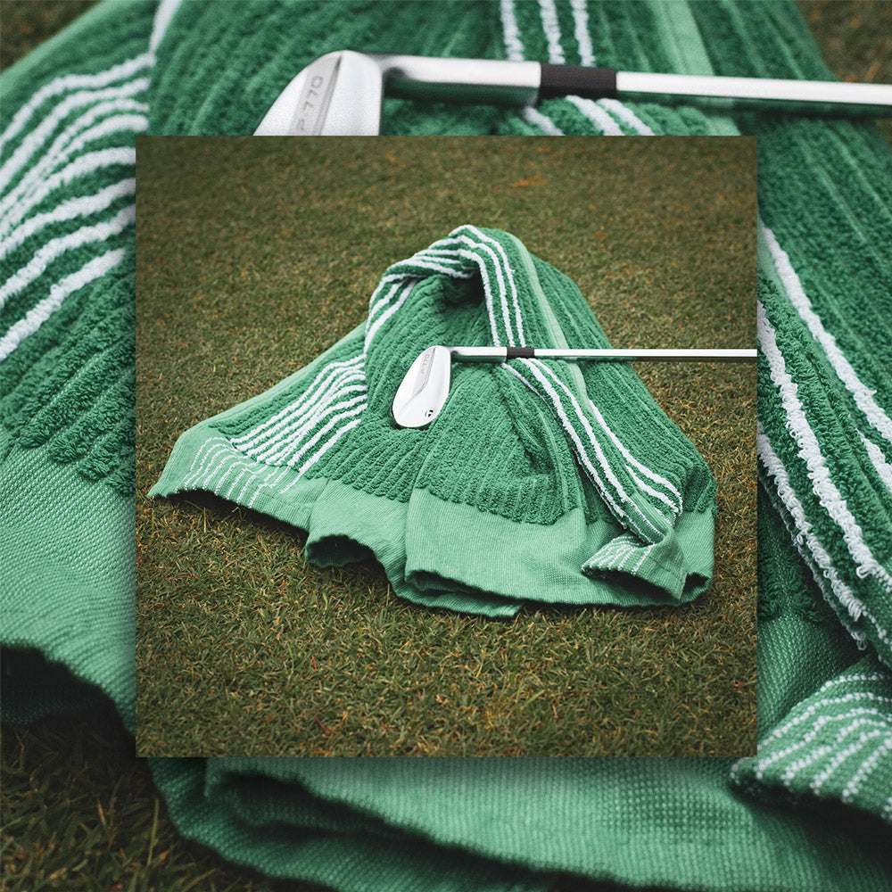 New green caddy towel from cayce golf laying on the fairway with an iron laying on top. The green caddy towel features white stripes. 
