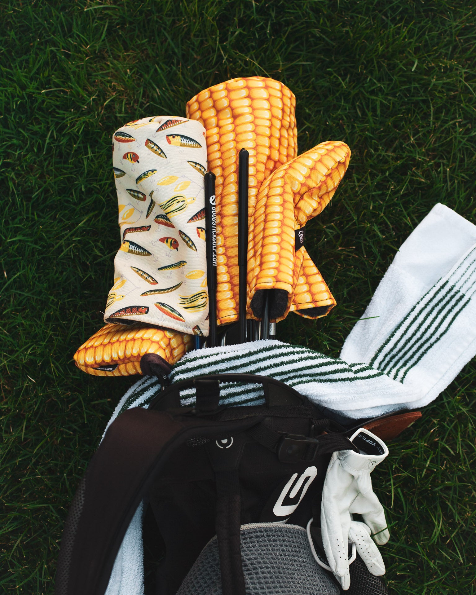 golf bag laying on the ground with corn golf head covers, fishing lures 3 wood headcover, and a green caddy towel